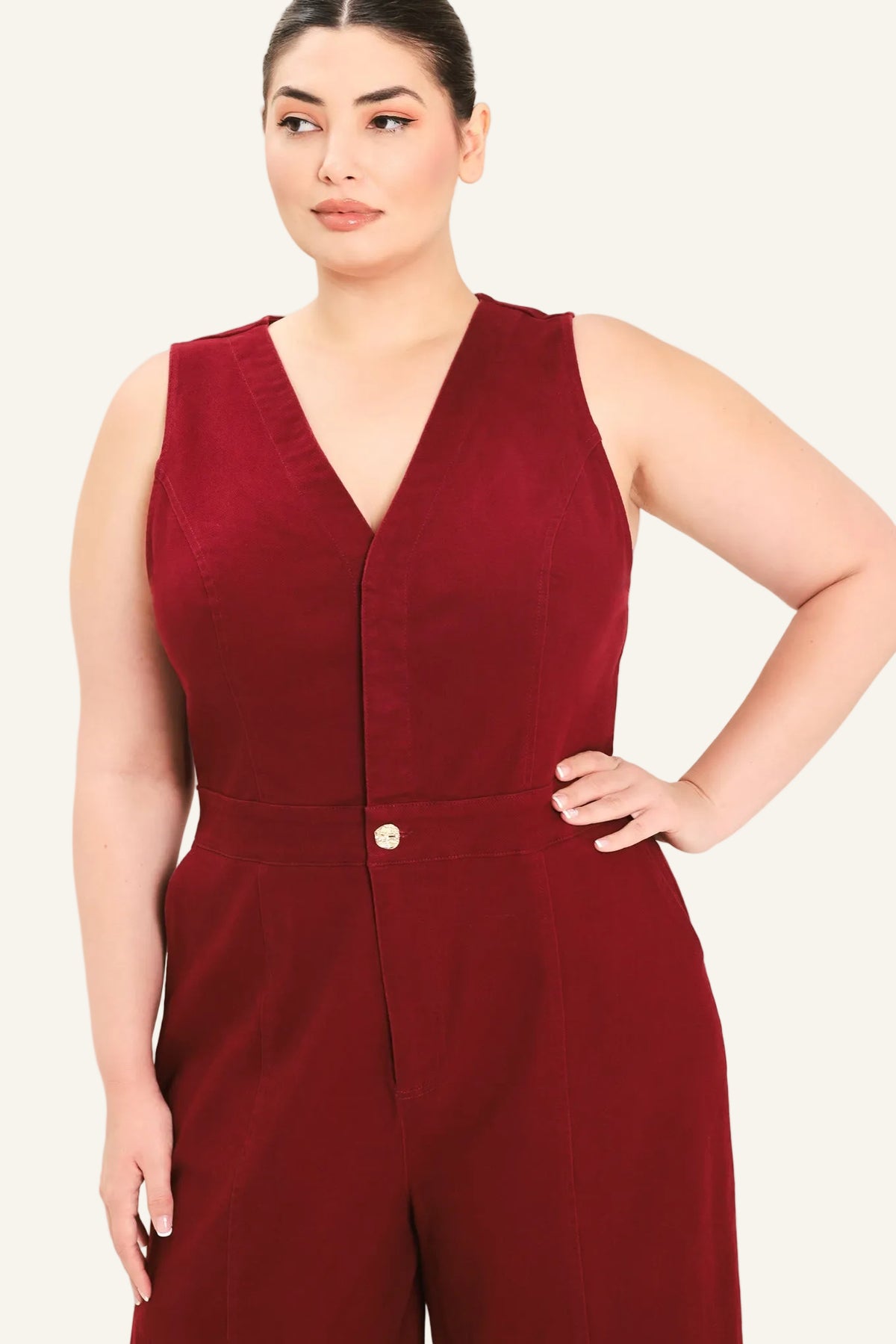 A woman modeling a sleeveless burgundy jumpsuit with a V-neckline and front button detail, photographed against a plain light beige studio background.