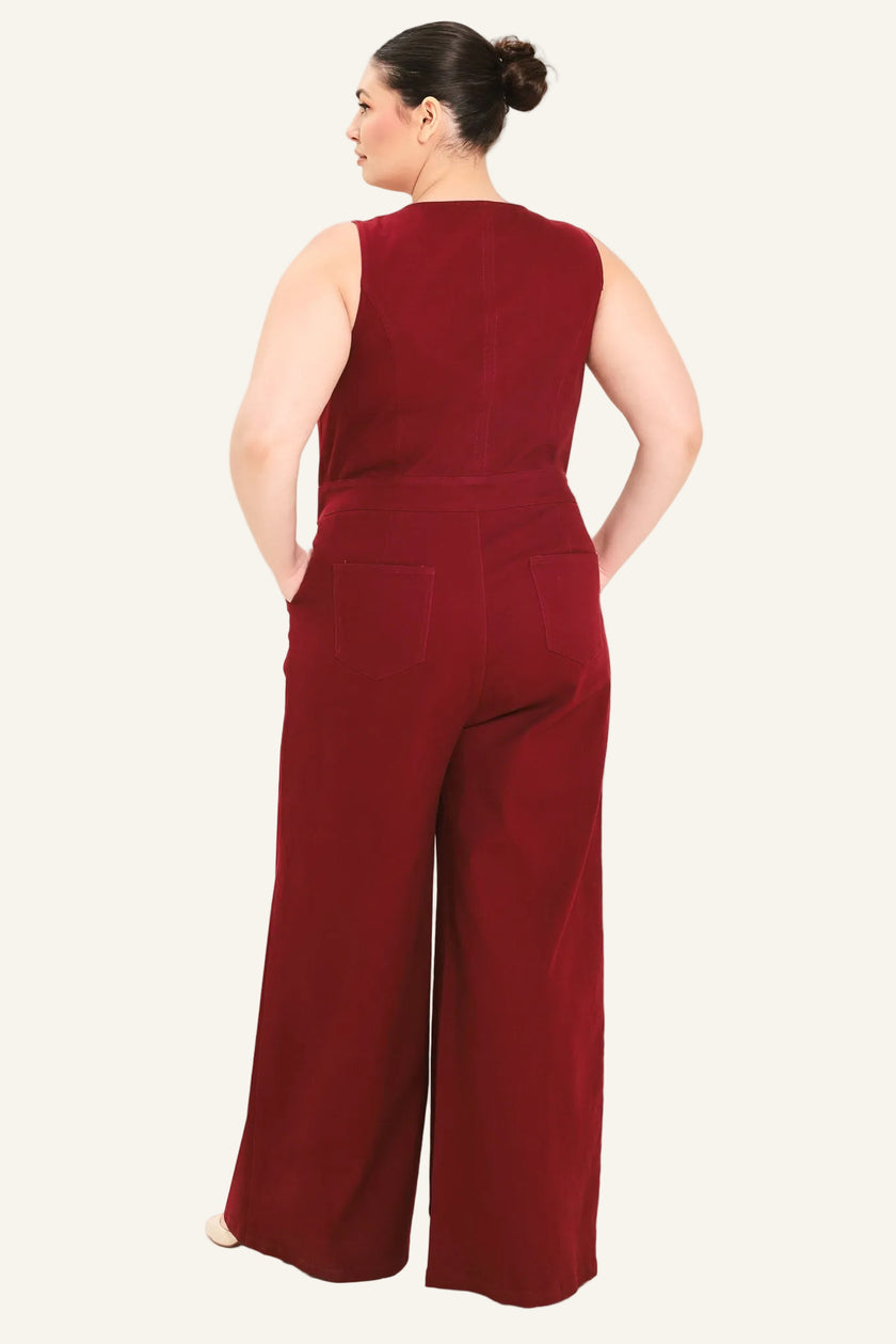 A woman shown from the back wearing a sleeveless wide-leg jumpsuit in deep burgundy, set against a plain light beige studio background.