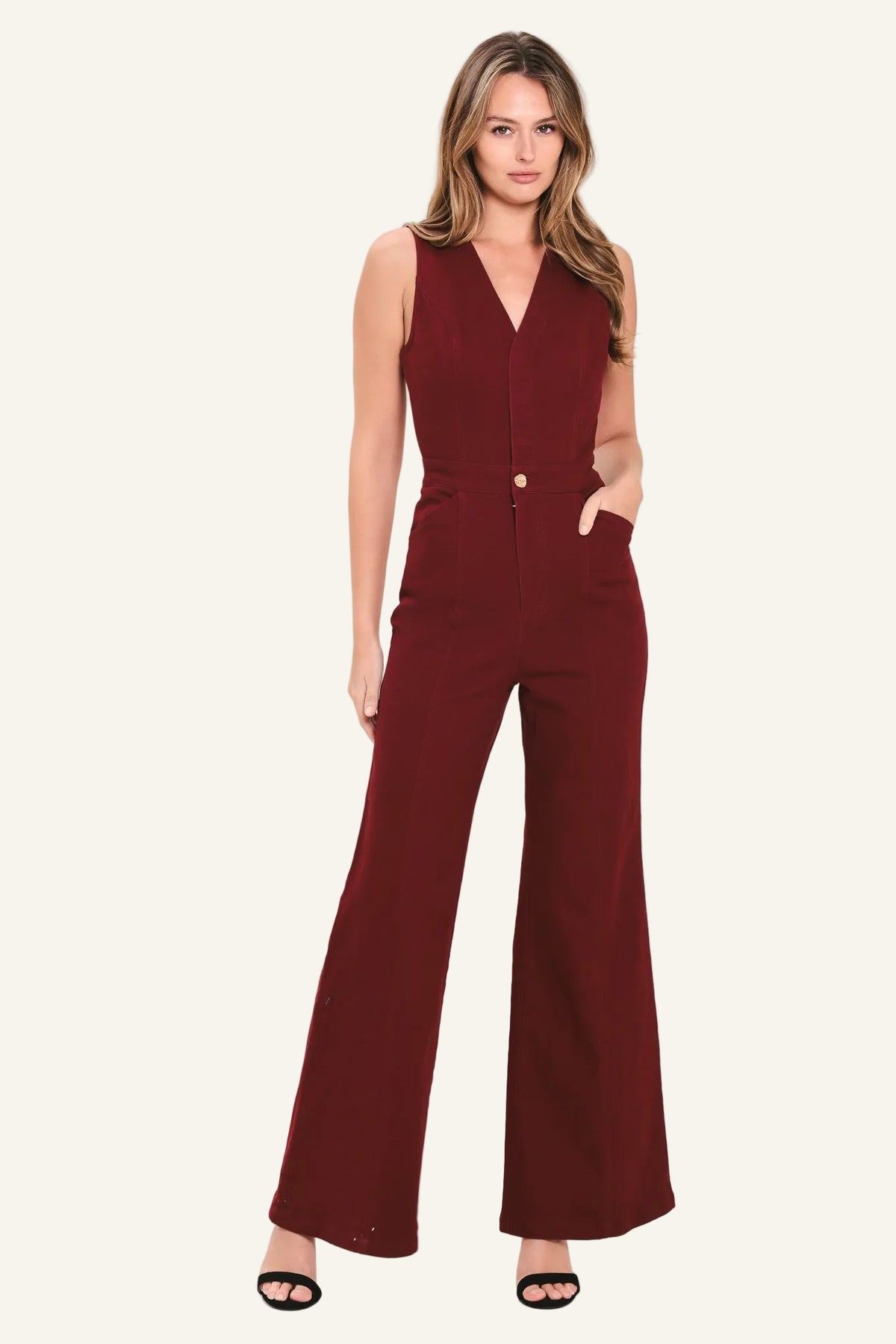 A woman modeling a sleeveless, deep burgundy jumpsuit with a V-neckline, front buttons, and wide-leg pants, set against a plain off-white studio background.